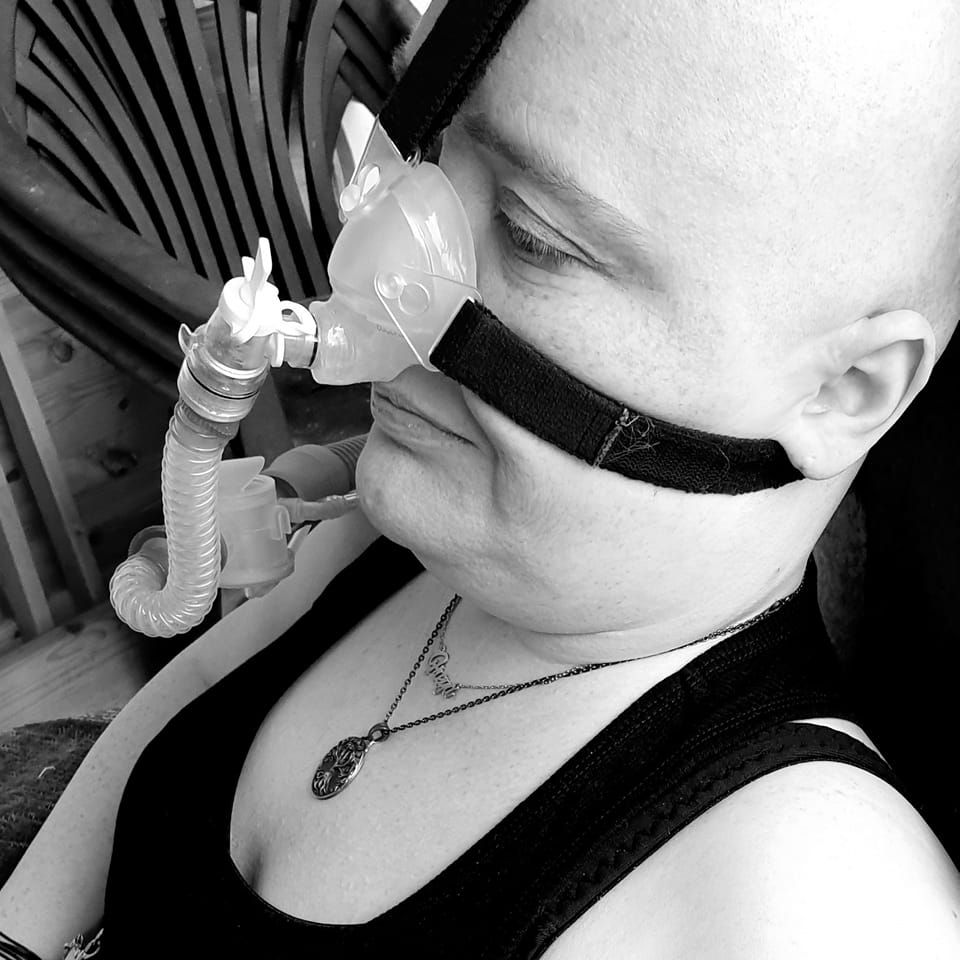 Das Schwarzweißfoto zeigt eine Person mit Glatze und einer CPAP-Maske im Gesicht, die an einer Schlauchverbindung befestigt ist. Die Person sitzt auf einem Stuhl und trägt ein schwarzes Top. Um ihren Hals trägt sie eine Halskette mit einem Anhänger. Der Ausdruck der Person ist ernst und nachdenklich. Der Fokus liegt auf dem Gesicht und der Maske. Der Hintergrund ist unscharf, aber ein Teil eines Gartenstuhls ist zu erkennen. Das Bild vermittelt einen Eindruck von Ruhe und Gelassenheit.