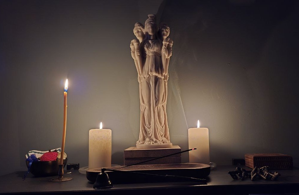 Das Bild zeigt einen kleinen Altar mit einer Statue der Göttin Hekate, flankiert von zwei brennenden Kerzen. Eine lange, brennende Bienenwachskerze steht auf der linken Seite. Vor der Statue befindet sich ein Räucherstäbchenbrenner mit einem brennenden Räucherstäbchen. Auf dem Altar befinden sich auch weitere kleine Gegenstände, darunter eine kleine Schale und eine kleine Holzkiste. Das Ambiente ist dunkel und stimmungsvoll. Das Bild wirkt meditativ und spirituell.